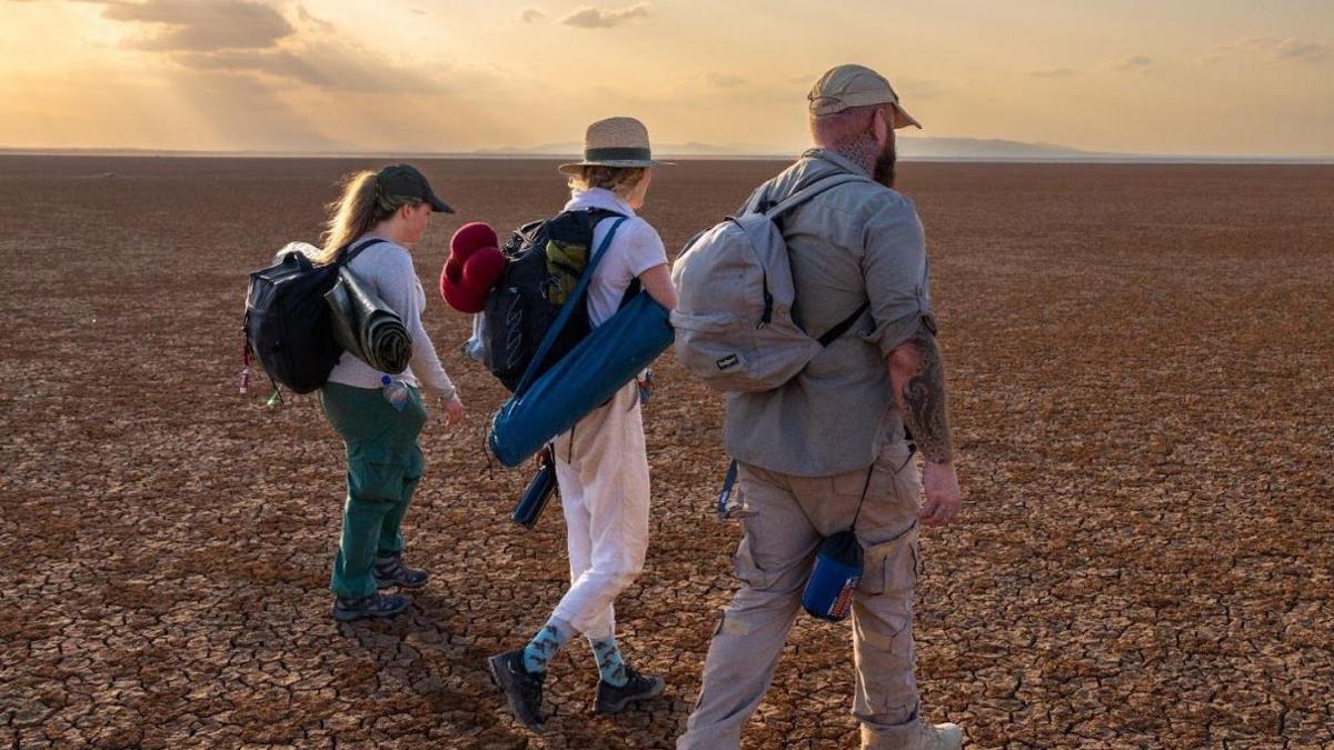 Three people (one man, two women) walking across a North African desert for the Channel 4 TV show Go Back To Where You came From - clouds and golden sky in the distance, cracked earth on the ground