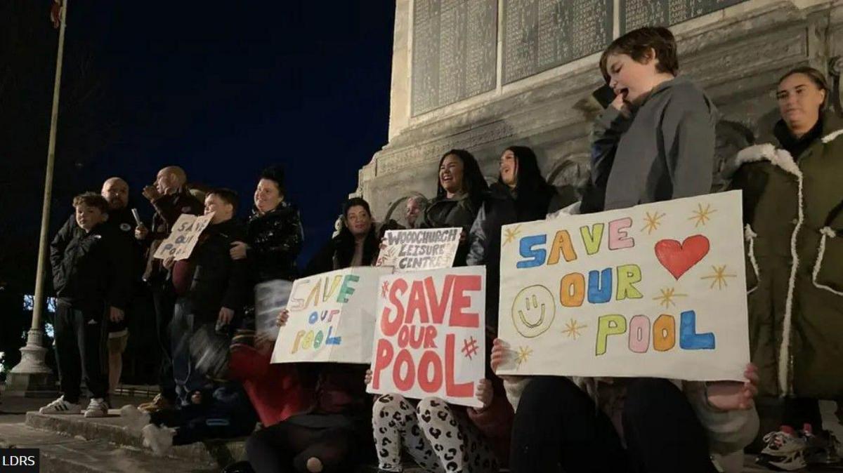Adults and children hold signs at a protest, including four which say "save our pool".