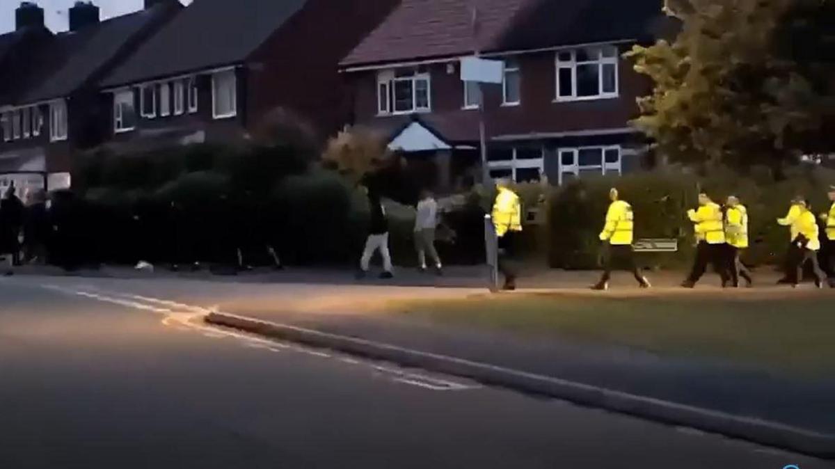 People dressed in black and police in yellow vests walking down a residential street. There are houses visible in the background