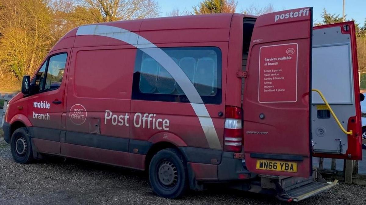 A red minibus that is a mobile post office. The outside of the van says "Mobile branch" and "Post Office" in white lettering. It also has the Post Office logo on it. The back door of the van is open