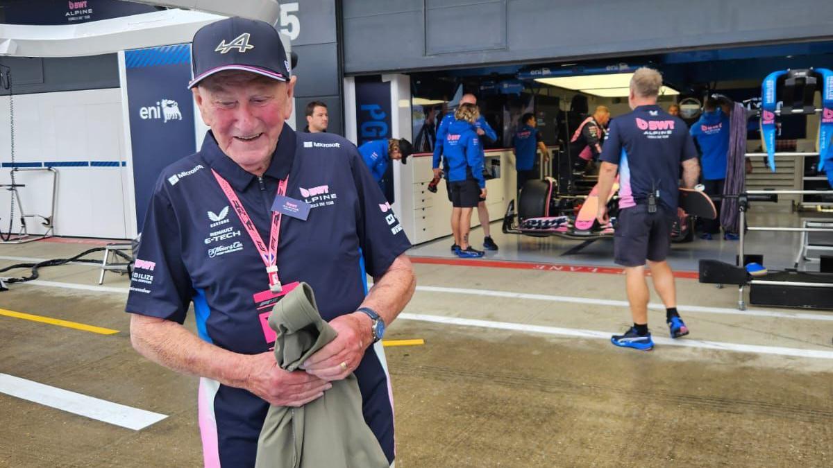 A man stands in front of a Formula 1. He is wearing a navy shirt with pink and white accents and holding a jacket. Team members are visible inside the garage, and the ground appears wet.