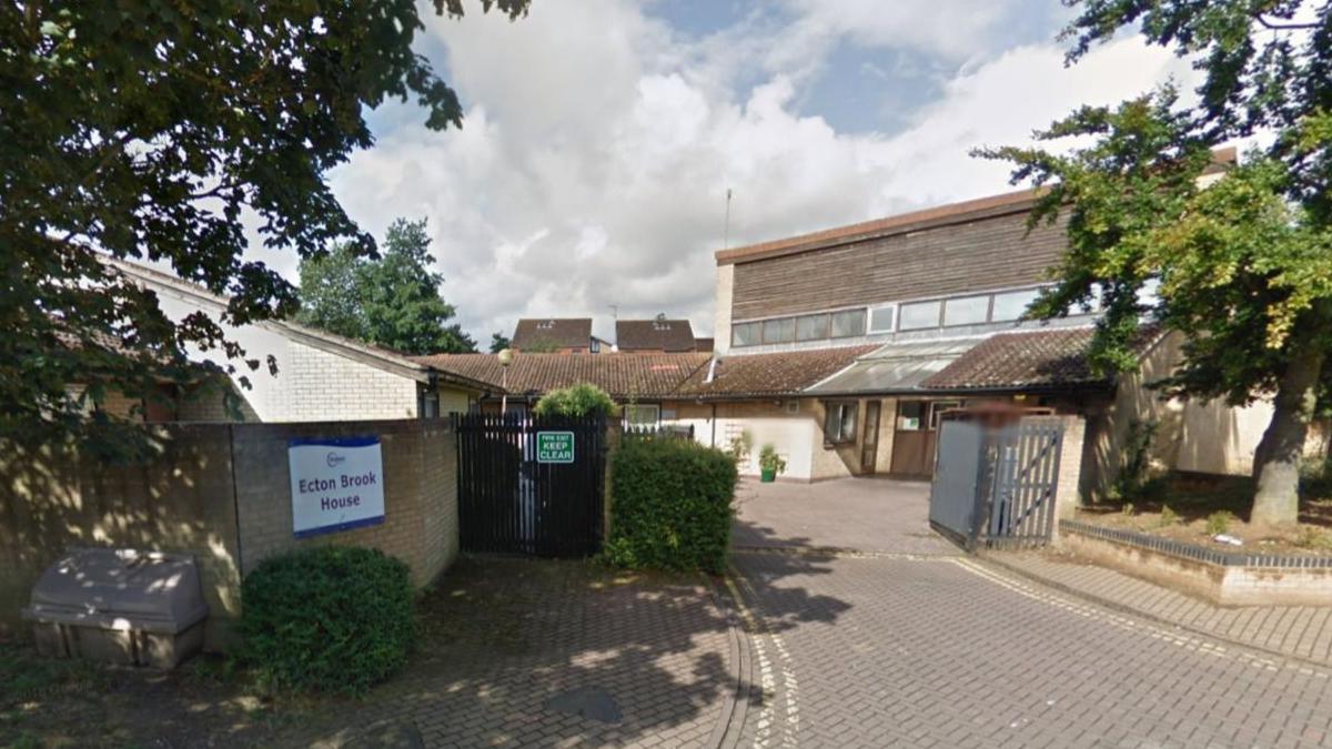 A brick and wooden-clad two-storey building standing inside a gated driveway. There is a sign on the wall saying "Ecton Brook House"