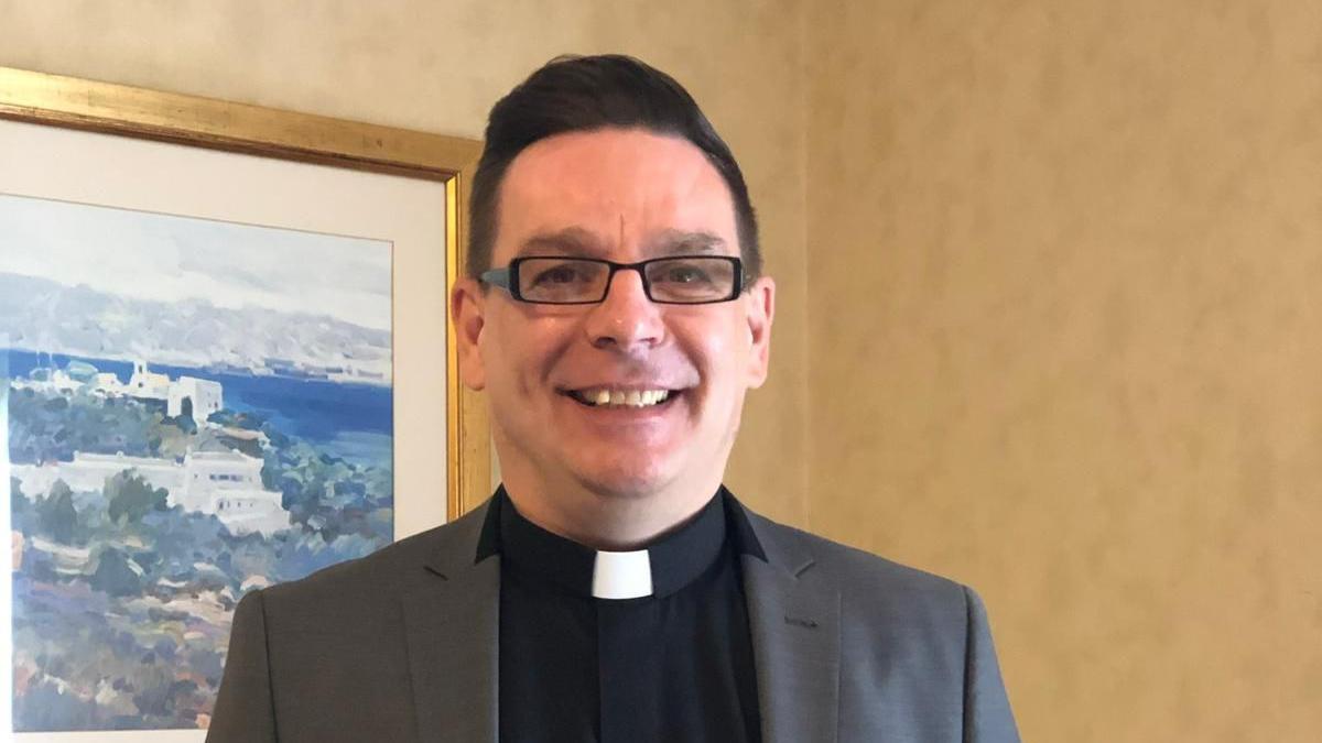 A man wearing rectangle glasses smiles at the camera, the picture is cropped to just his head and shoulders but you can see he is wearing a clerical collar - better known as a dog collar - and a grey blazer. The background shows yellow wallpaper and a painting of a Greek town.