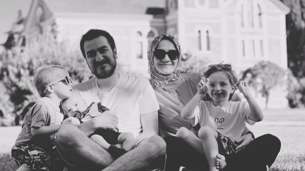 Zehra Gültekin and her husband Bilal Gültekin smile in a black and white photo seated with their children on a lawn in front of a large building. Zehra is wearing a headscarf and dark glasses, and one child - on the far left of the picture - is also wearing sunglasses