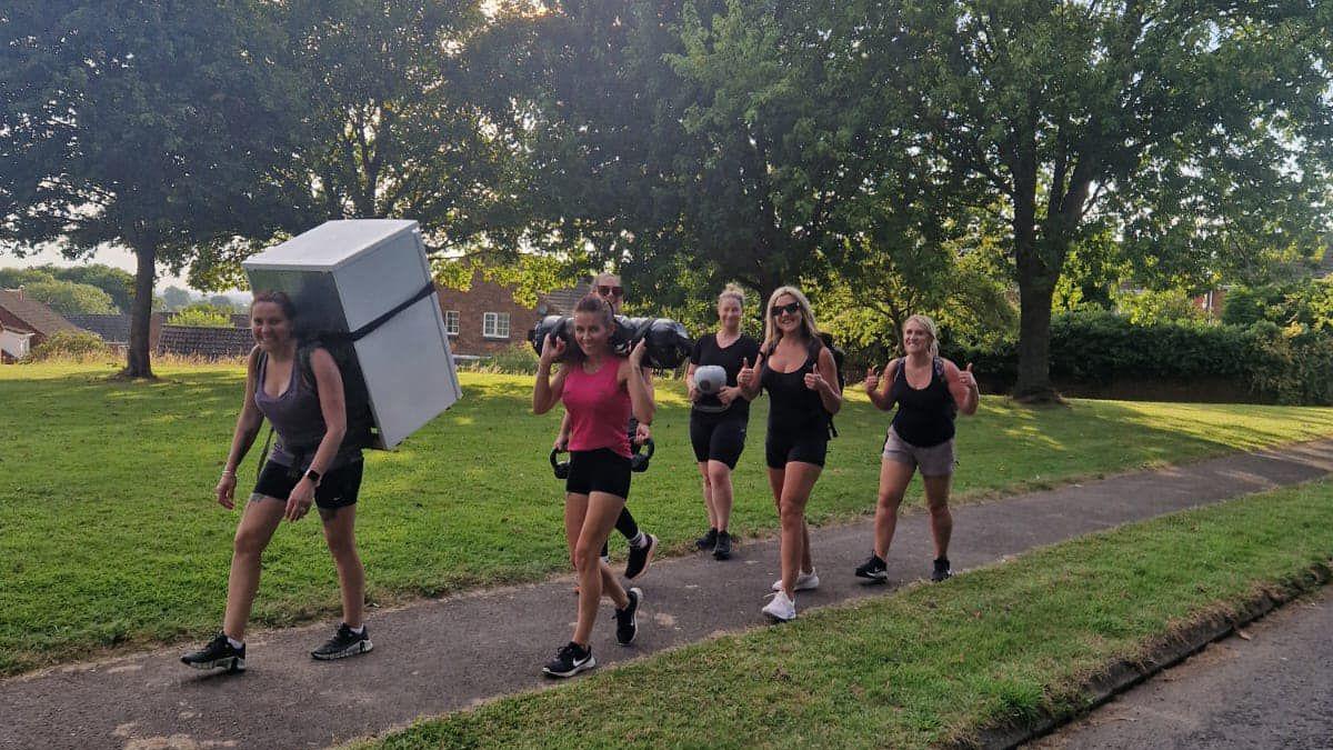 A group of six women walking through a grassy area carrying a fridge and weights