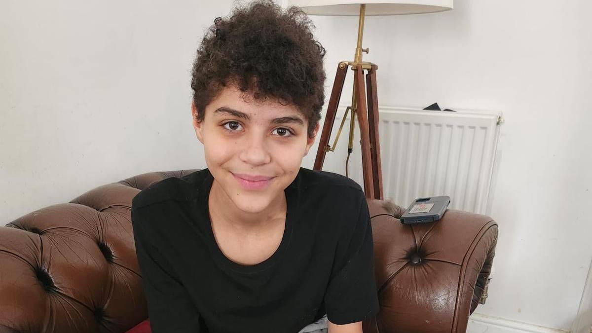 A 16-year-old boy called Zak is smiling looking at the camera, sitting on a brown sofa in his living room.