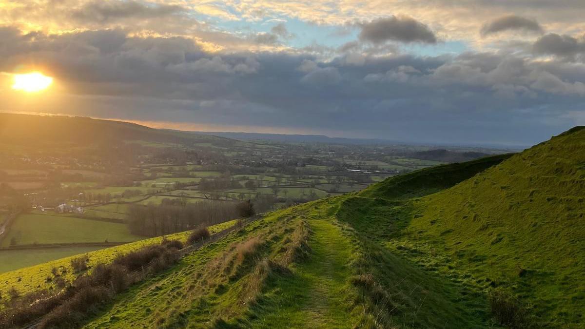 A panoramic view of the countryside with a patchwork of fields and trees stretching to the horizon. In the foreground a path can be seen winding round the side of a hill covered in green grass. The sun is setting casting a golden glow across the left-hand side of the shot.