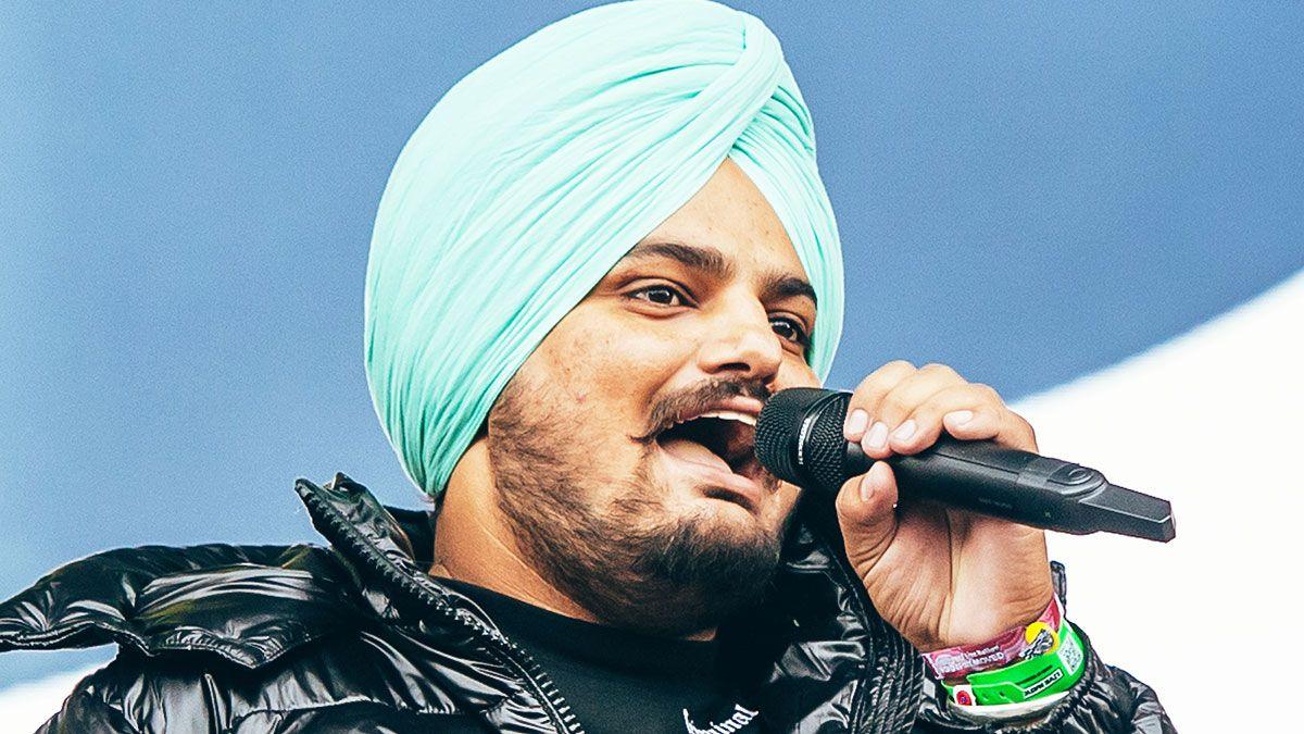 Moose Wala is wearing a black puffa jacket, turquoise turban and is singing into a microphone.