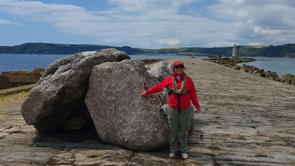 Jennifer Kloester stood in front of large rocks on the Plymouth breakwater. She is wearing a red jacket, a red cap and dark green trousers. In the distance, at the end of the long stone breakwater, stands the grey lighthouse. Surrounding the long pathway is the blue sea and in the distance green land. She is smiling at the camera with her left hand touching one of the large rocks.