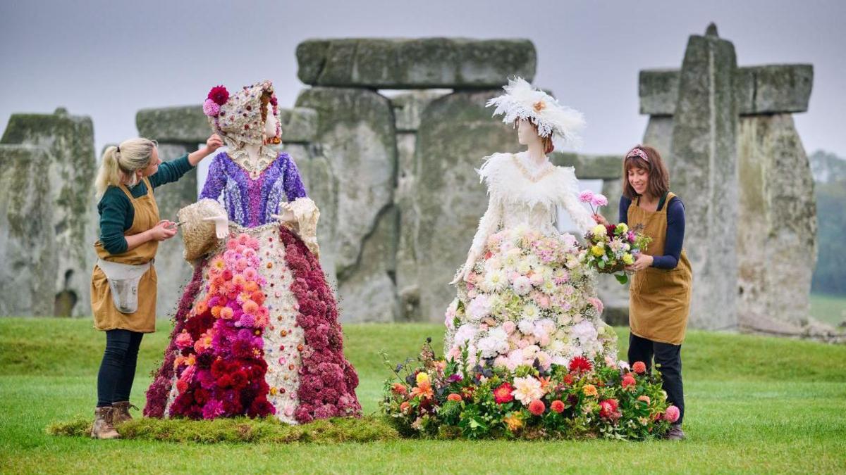 Two florists wearing brown aprons, delicately placing dahlias into two sculptures which depict Victorian women in hoop skirts. They are standing on the grass in front of Stonehenge, which can be seen in the background on a misty day.