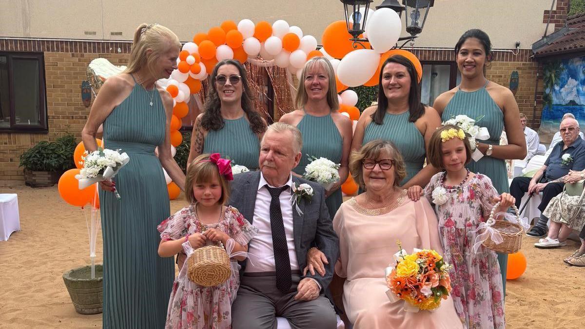 A wedding party sit in two rows for a photo, with the bride and groom in the front row with two flower girls and the bridesmaids behind. The groom has a light grey suit and dark tie on, and the bride has a light pink dress on. The bridesmaids are all in dark grey dresses.