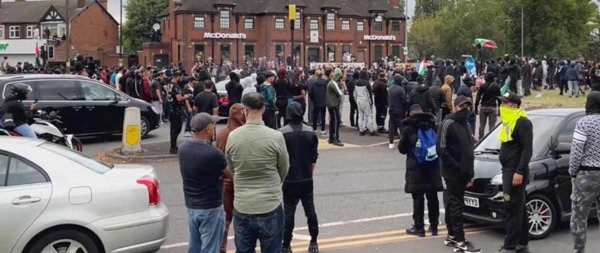 Scores of men are gathered in the middle of a street, amid cars, with their backs to the camera; many of whom are wearing face coverings.