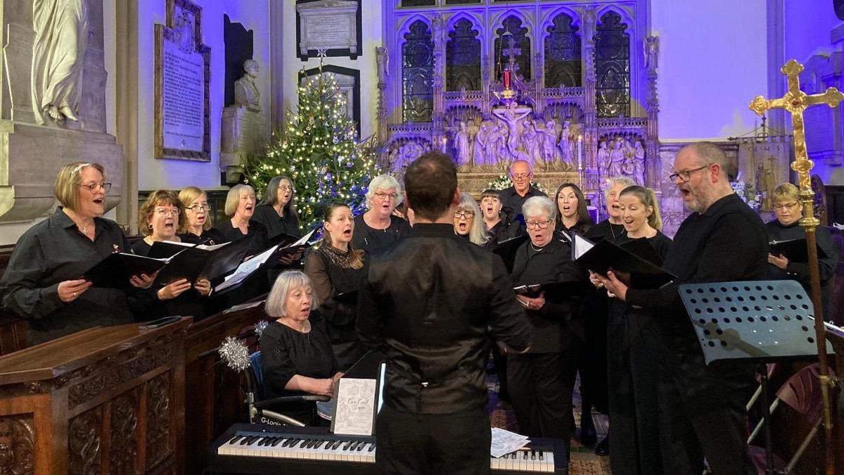 A choir wearing black clothing, shirts and trousers, sing in front of a man conducting them next to a piano synthesiser, who has his back to us. Behind them is a stone carving of a Biblical scene with a Christmas tree, covered in lights, to one side.