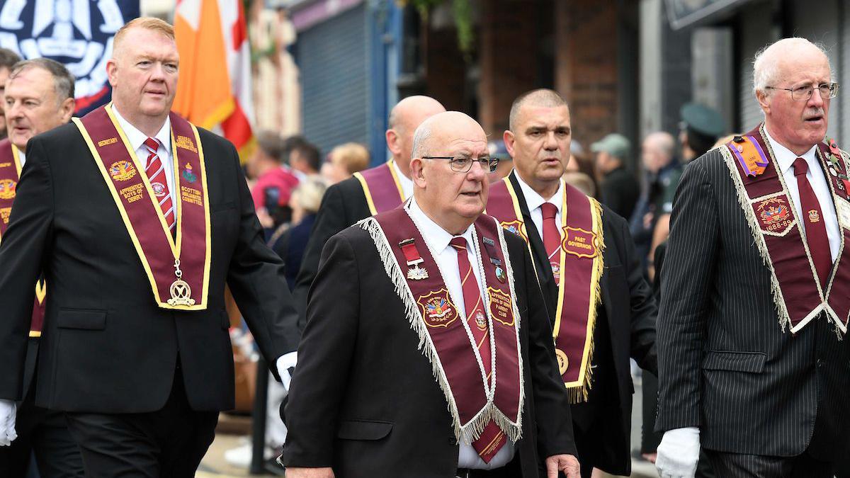 A number of men wearing suits with red ties, and ceremonial garments, marching in a parade.