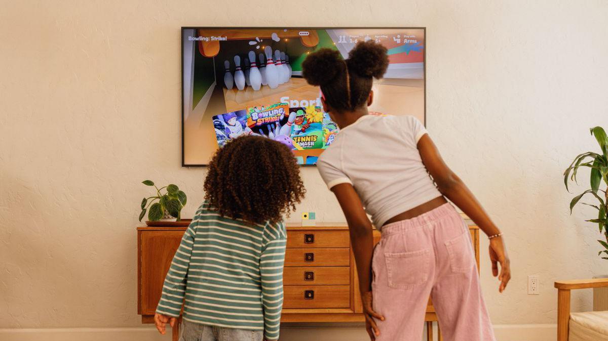 Two children are staring with their backs to the camera in a living room space facing the TV screen which is showing a bowling game. Beneath the screen the Nex Playground console cube in white, green and yellow sits on a table.