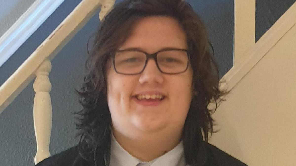 A teenage boy. He has long brown hair and black glasses.