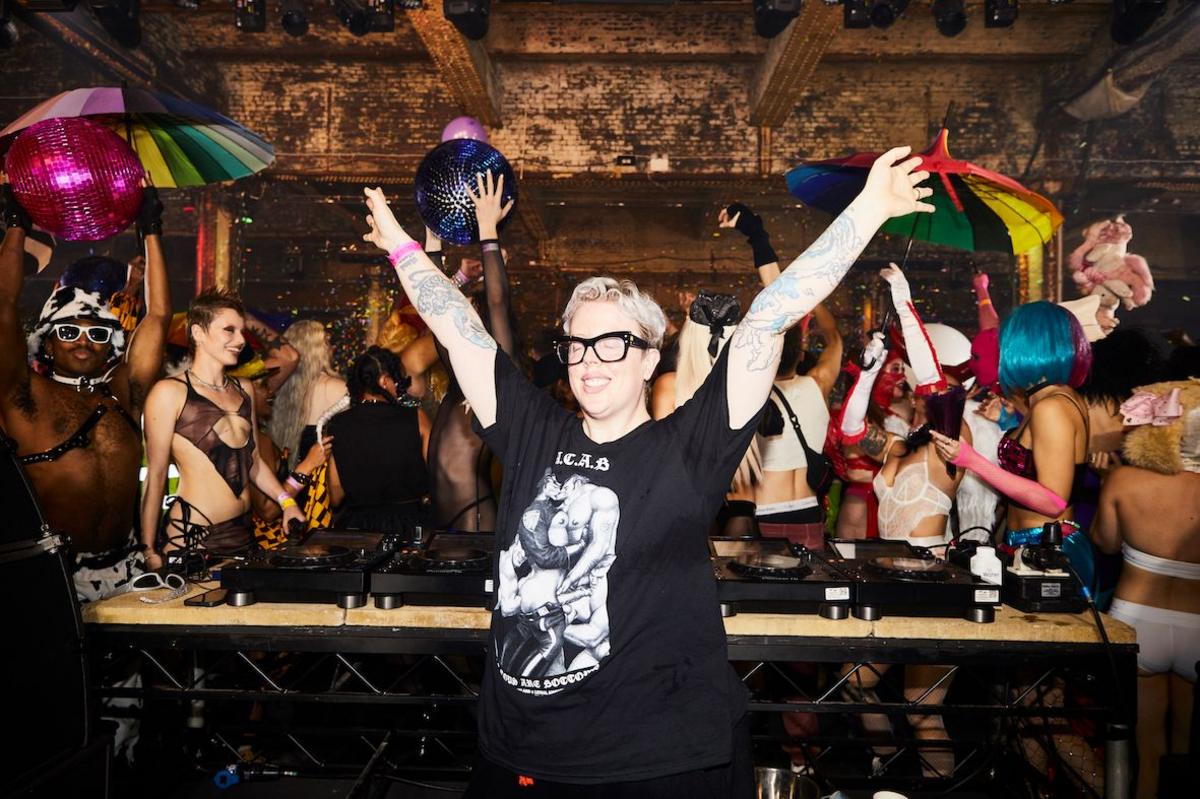 The Blessed Madonna poses with her arms in the air behind her DJ decks, while a colourful group of ravers dance behind her.