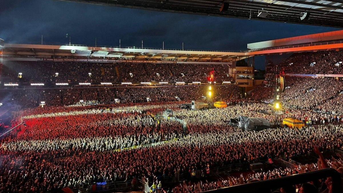 A huge sea of people are seen in the stadium, lit up by red lights from the stage
