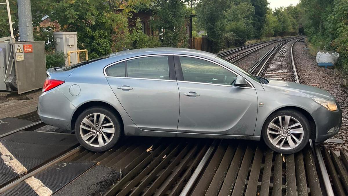 A silver car stopped on a level crossing with the tracks in the background, lined with trees.