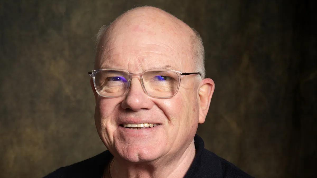Bald man with glasses smiling at the camera on a dark background.