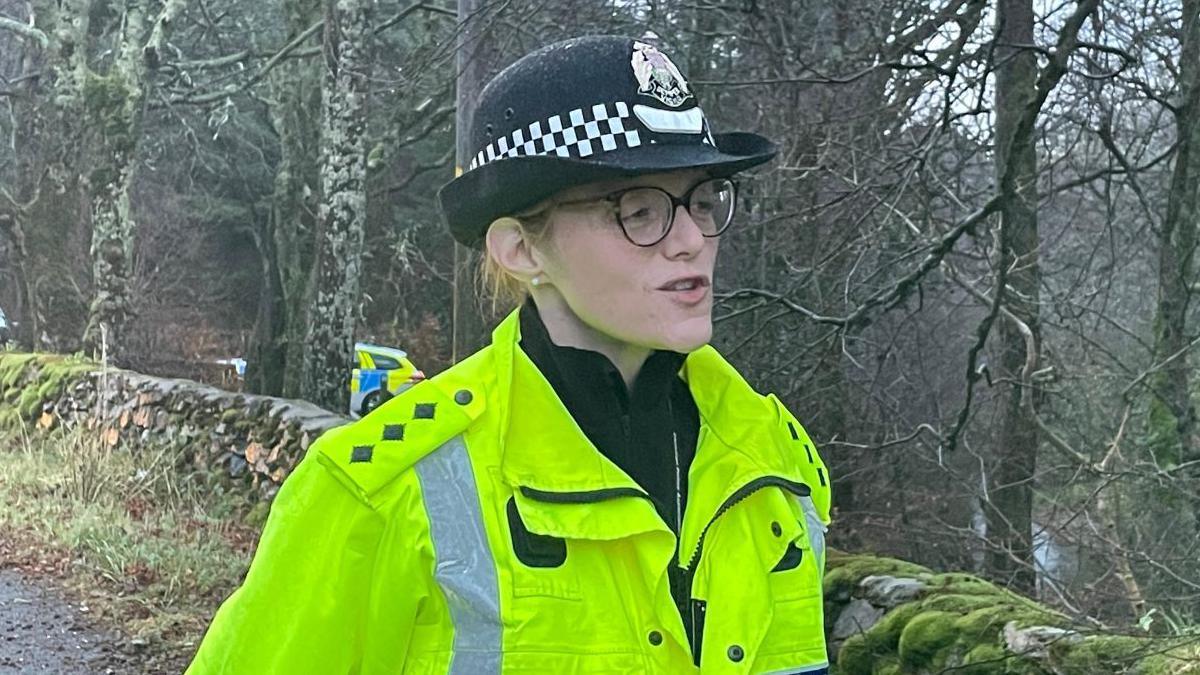 The police officer, who is wearing a police hat, fluorescent green jacket and a pair of glasses, speaks while standing near a mossy, stone wall and line of trees.