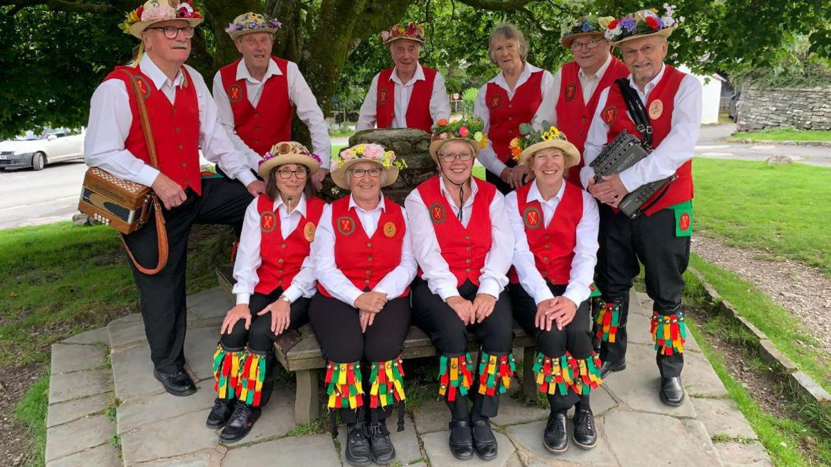 Four members of Furness Morris Dancers sat on a bench with six more standing behind them. They are all wearing black trousers, white shirts and red waistcoats. All but one have hats with flowers at the top. They are smiling. The phot was taken in 2023.