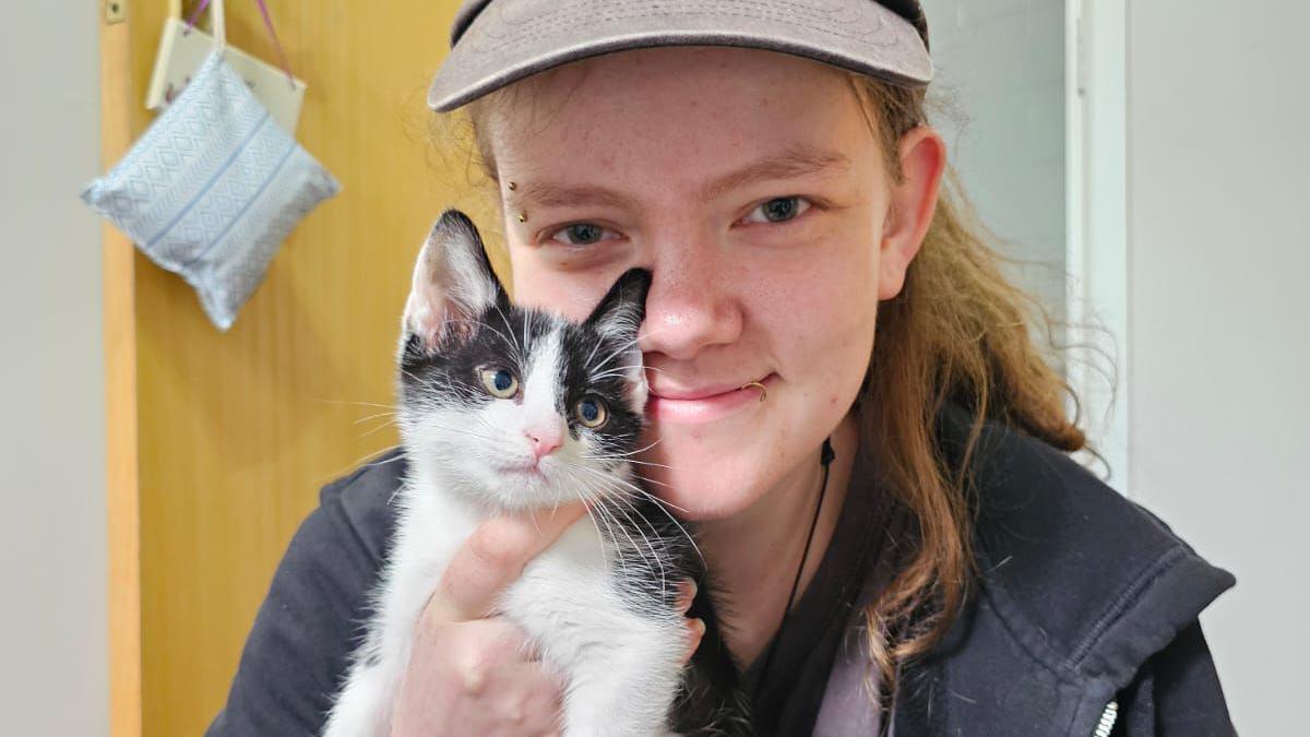 Chloe White, a woman wearing a cap and a navy hoodie with eyebrow and lip piercings, smiles while holding a black and white kitten.