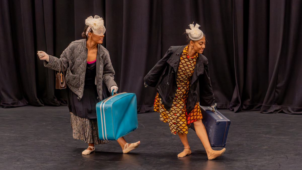 Two black women dancing on stage in front of a curtain. One is dressed in a colourful African print and the other is wearing a long dress and padded jacket. They are both wearing beige fascinators in their hair and carrying suitcases.