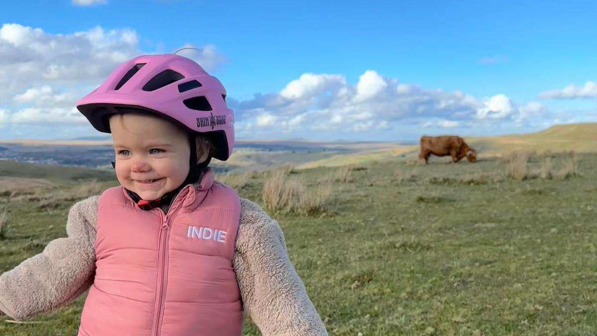 A little toddler wearing a pink helmet, a fleece and pink body-warmer with her name 'Indie' on it in white writing. She's on a mountain with a brown highland cow in the background. The sky is bright blue with some white clouds.  