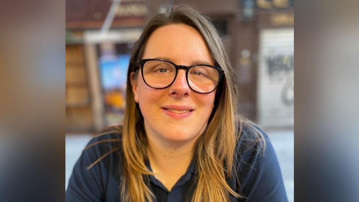 Llun bortread o Meg John, Rheolwr Cefnogi Digwyddiadau ar gyfer elusen Pride Cymru, yn eistedd gyda'i breichiau wedi'u croesi o'i blaen. Mae ganddi sbectol am ei thrwyn a gwallt hir brown golau.