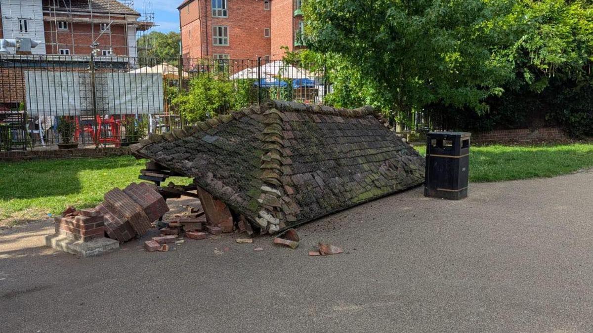 Bricks and a large tile roof of a historic outdoor shelter. The structure has collapsed next to a black public waste win on a concrete surface. In the background are trees and grass, with residential properties beyond them.
