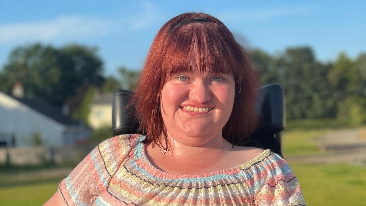 A woman, pictured from the chest up, smiling at the camera. She is wearing a stripy orange, yellow, white and blue top. She has dark red hair. She is sitting in a wheelchair. The background of the photo is blurred with grass on the ground and a blue sky above. 