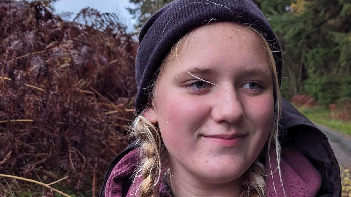 Amelie wearing a dark coloured beanie hat with blonde hair in braids. she is wearing a rain mac over a mauve hoodie and is standing outside on a path.