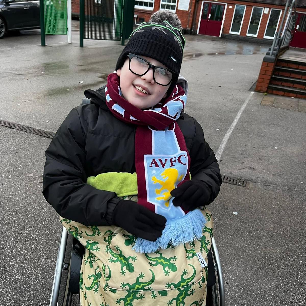 A boy smiles at the camera while sitting in his wheelchair. He has a maroon and blue Aston Villa scarf wrapped around his neck and has a yellow blanket with green lizards on it around his lap. He wears a black coat and hat and is sat outside on tarmac, in front of a redbrick building.