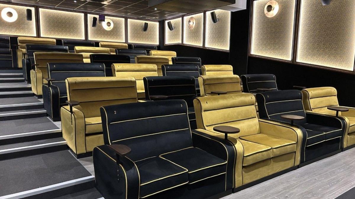 The inside of a cinema with black and gold two-seater sofa-style chairs. Stairs are on the left and there are little tables in between each two-seater.