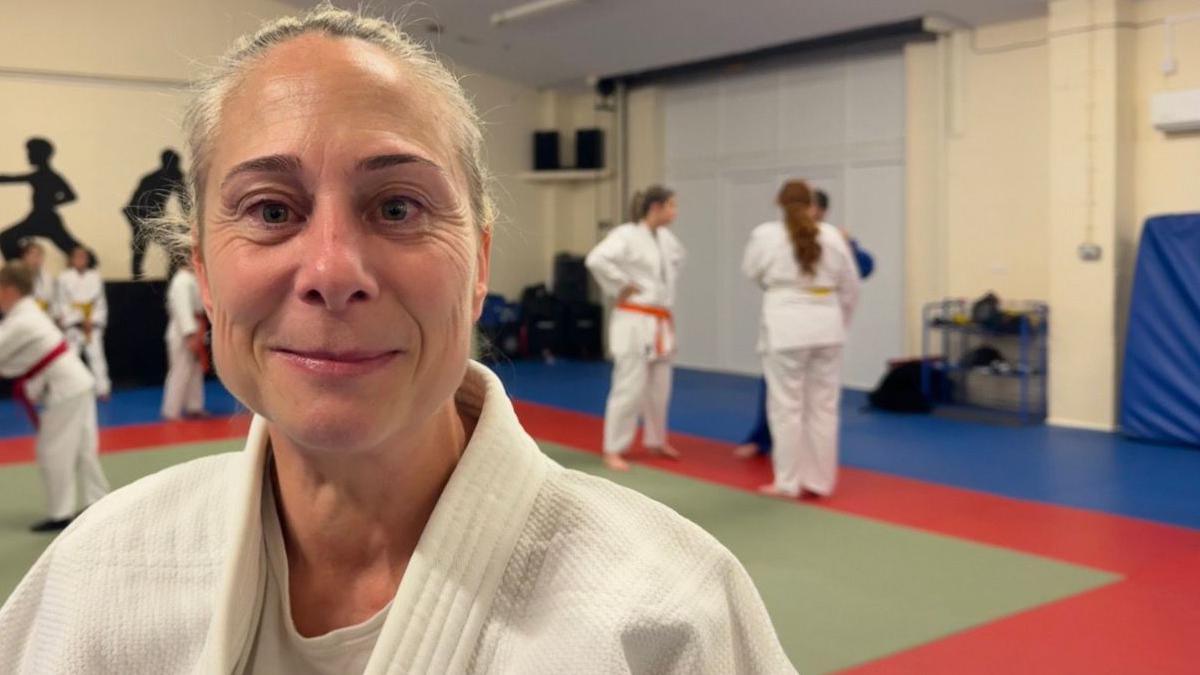 A woman looking at the camera, her face set in a half smile. She has her hair pulled back into a ponytail. She's wearing a white judo jacket. In the background there are children who are also wearing white jackets, and are taking part in a training session. The walls are decorate with large silhouettes of people in judo poses.
