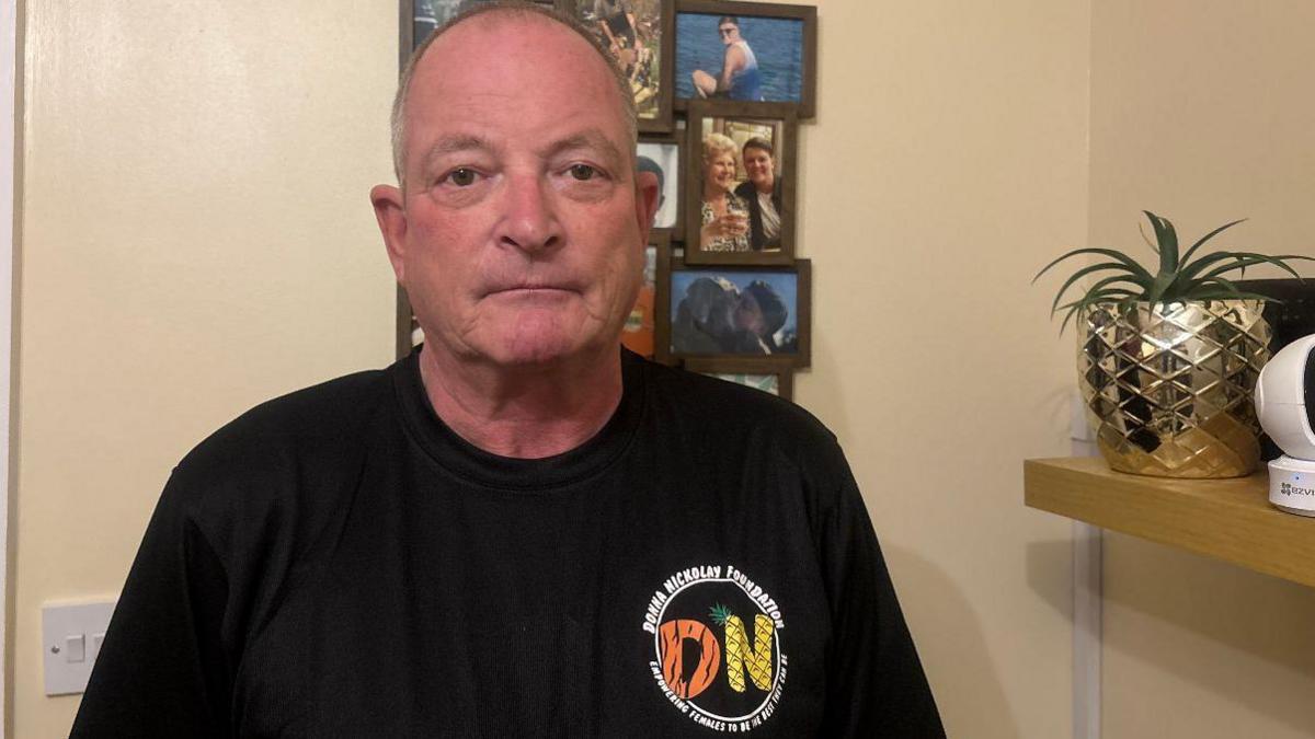 A man with a glum face, wearing a black shirt with the Donna Nickolay Foundation logo on. He is stood behind a series of pictures of his late daughter Donna inside his house