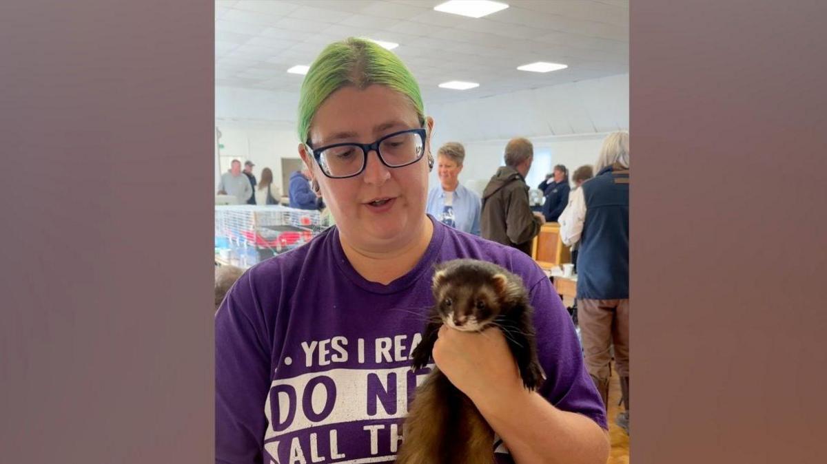 Heather Johnstone holding a brown ferret. She has green hair, glasses with a blue rim and a purple t-shirt.