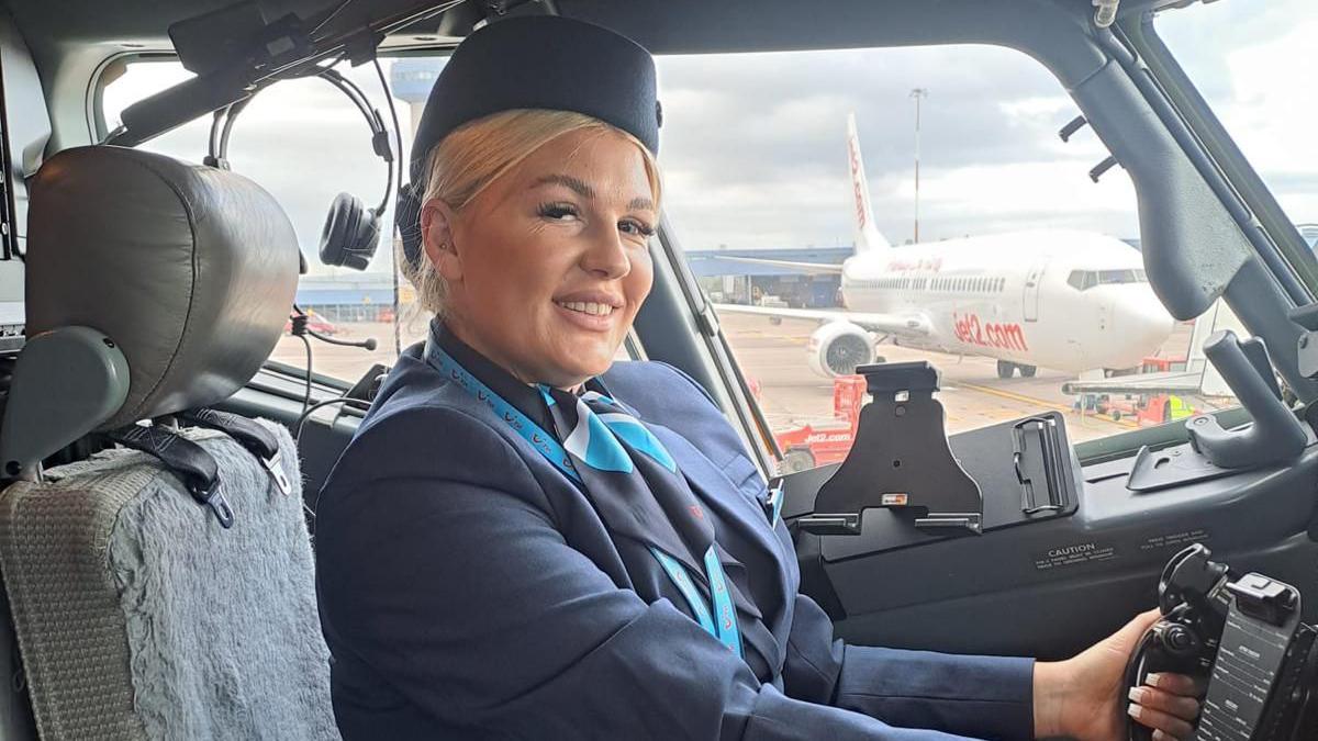 A woman in a navy flight attendant uniform poses at the controls of a plane