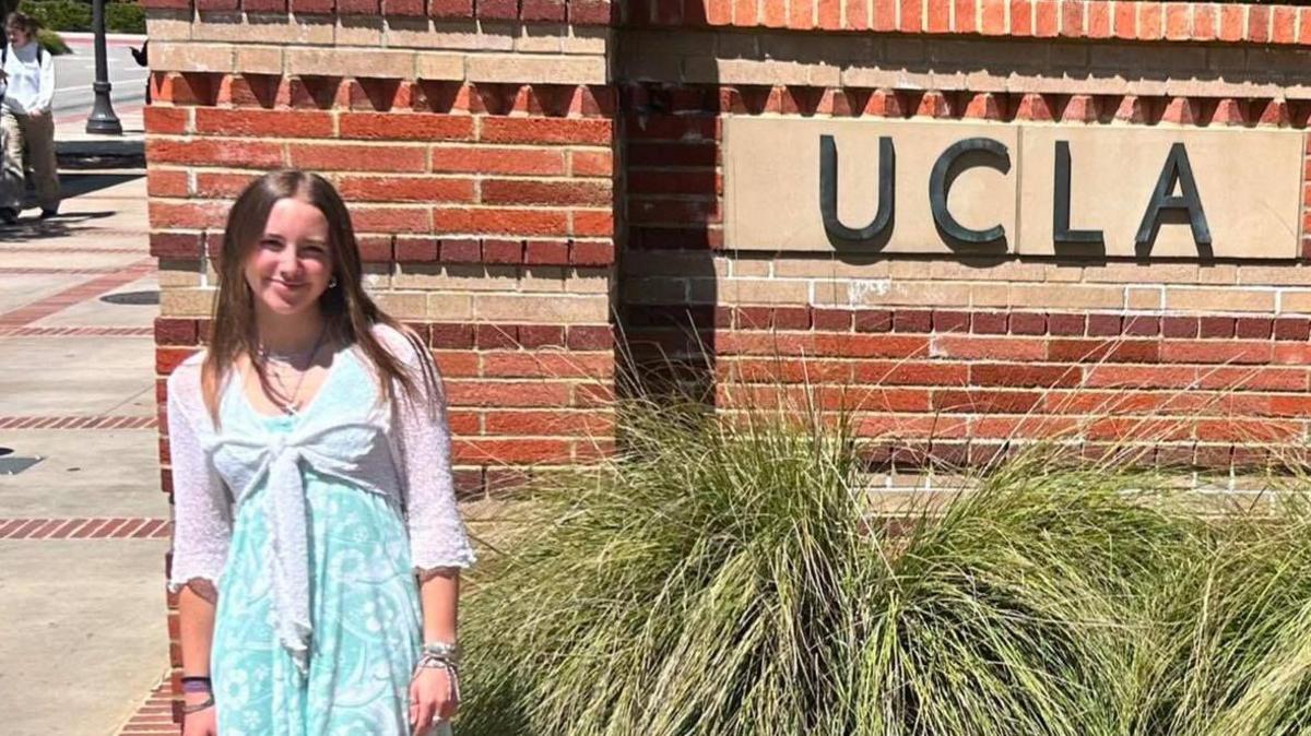 Amelie Le Prevost stands in front of a brick wall which has 'UCLA' written on it. There are tall grasses next to her. She is wearing a light blue dress with a white cardigan. 