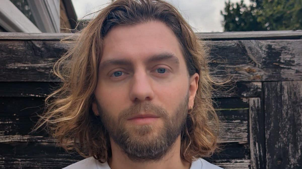 Luke Pickett appearing with long brown hair combed back and a beard standing in front of a wooden fence. His ears are pierced.