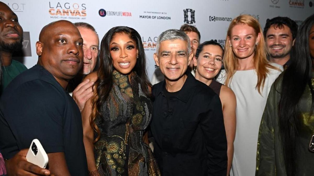 Sadiq Khan stands in the middle of a group of people posing for a photo. He is wearing a black t-shirt and black jacket.