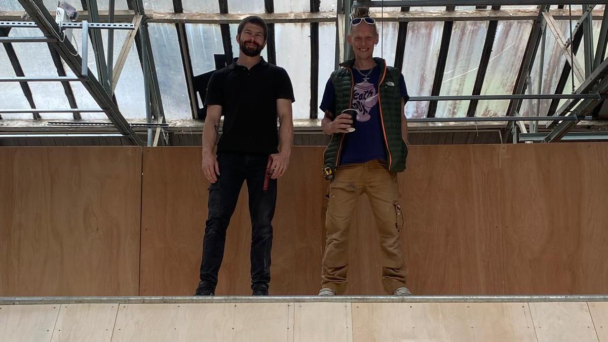 Two men stand at the top of a wooden skateboard ramp, looking down at the camera. The man on the left has short dark hair and a dark beard and is wearing jeans and a dark polo shirt. The man on the right is wearing brown trousers and a blue t-shirt under a green sleeveless jacket. He is holding a takeaway coffee cup and has a pair of sunglasses perched on his head.