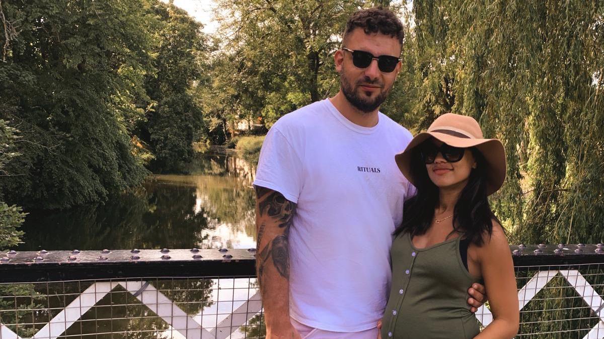 Charlotte Lescott stands next to her partner Ali Watson. Charlotte is pregnant. They are on a bridge which goes over a river.