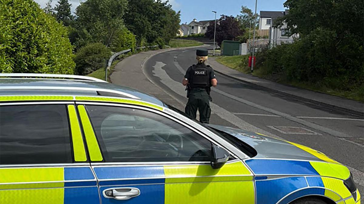 A police officer is standing in front of a police car. They are wearing full uniform including a hat. The background is a black road, footpath and crash barrier. There are houses and trees visible.