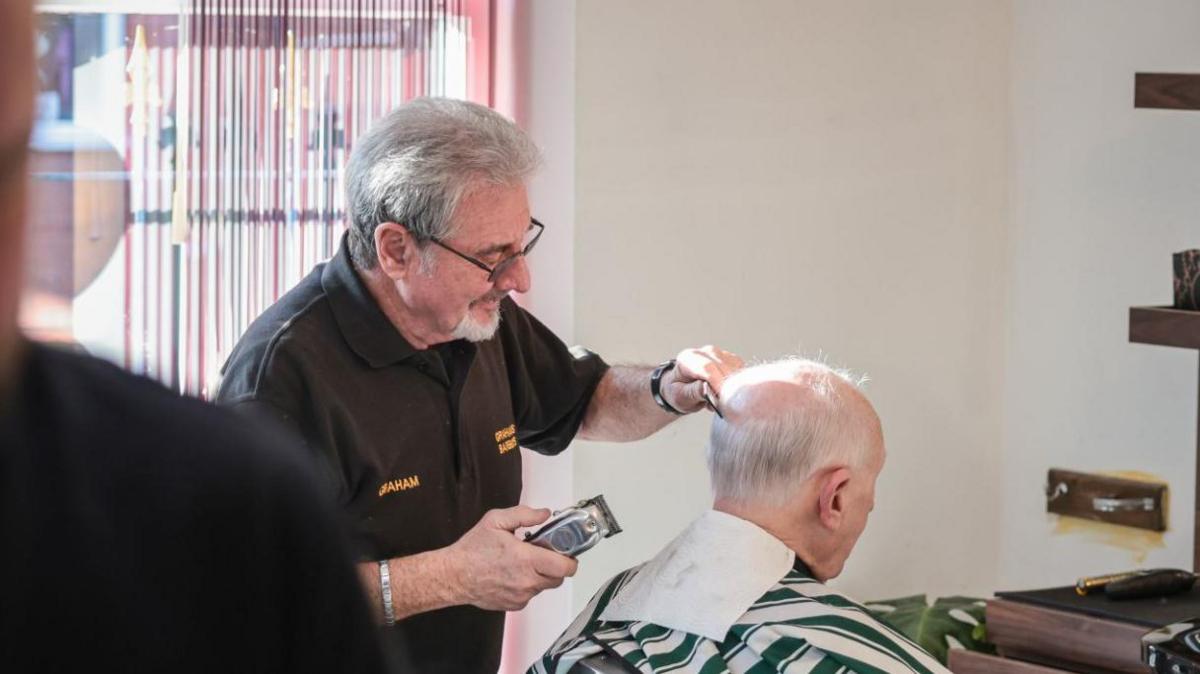 A man wearing a brown short-sleeve polo shirt holds a pair of hair clippers in one hand and a small comb in the other as he prepares to cut a man's hair