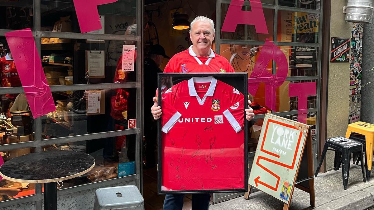 Mike stood outside the pub holding a framed Wrexham shirt which has been signed.