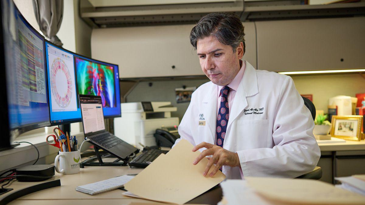 Man in a pink shirt and tie with a white lab coat flips through medical reports in his office. 