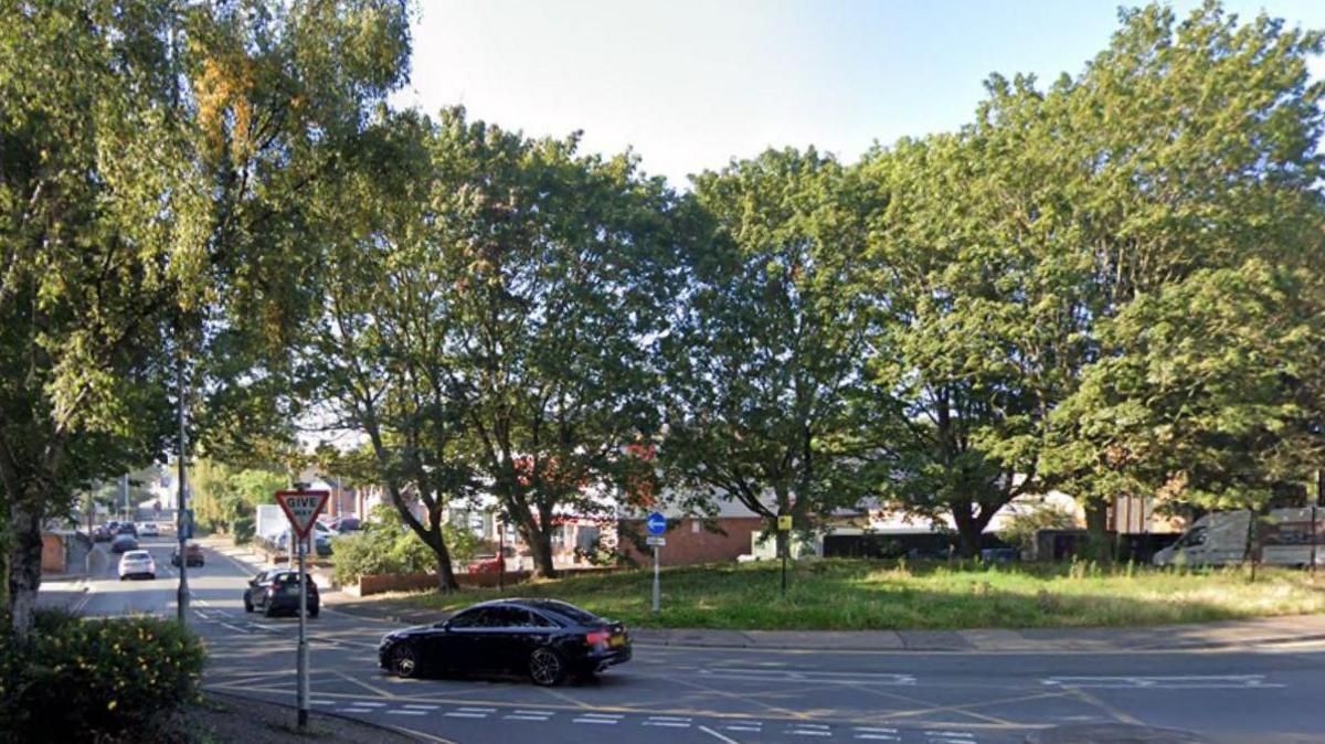 A car is driving around a roundabout with grass and tall trees in the centre and a yellow box junction in the foreground.  