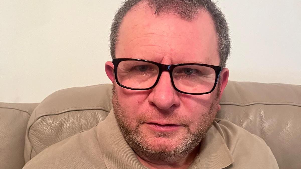 A man in black glasses and a beige t-shirt sitting on a beige leather sofa. A cream wall is behind him.